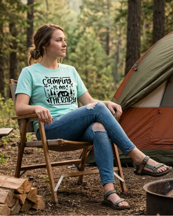 Woman sitting in foldable camp chair next to a tent and wearing a t-shirt that says camping is my therapy