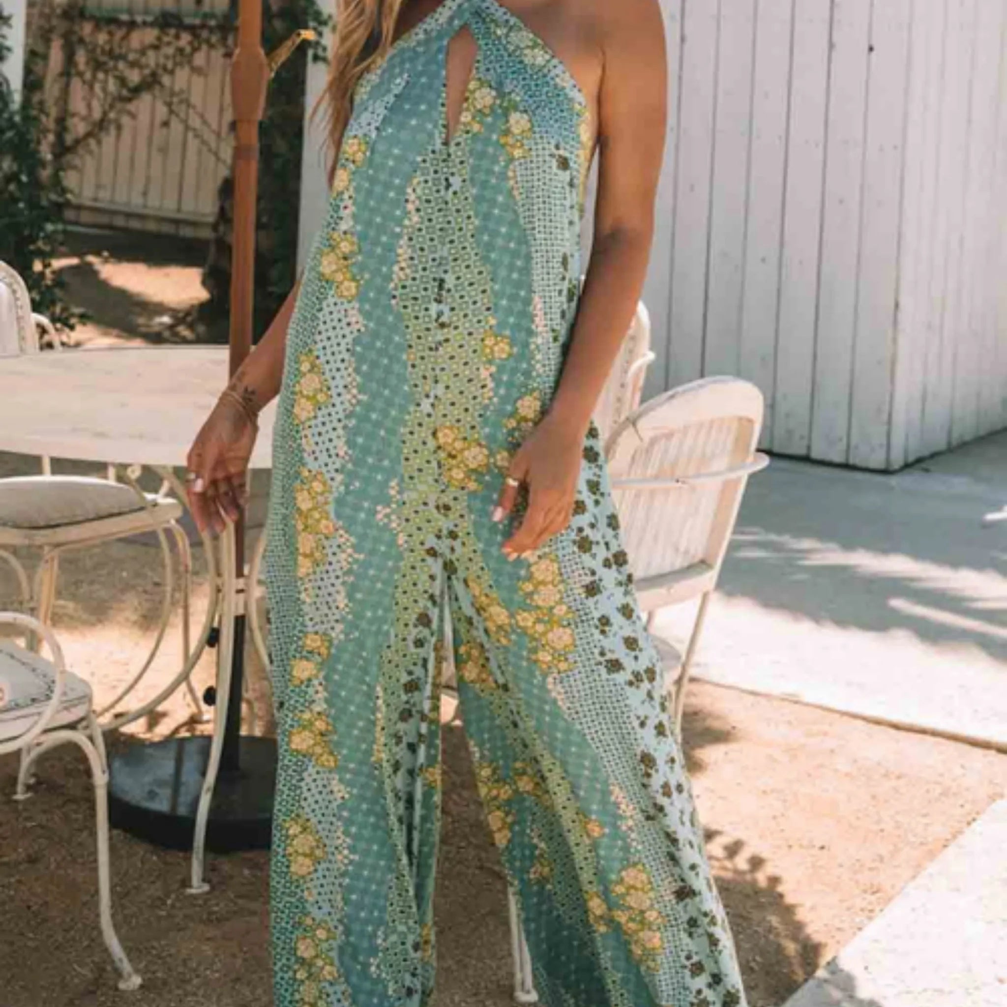 Woman wearing a floral dress standing outdoors with chairs and a table in the background.