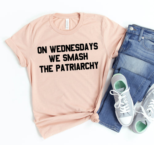 Pink t-shirt with 'On Wednesdays We Smash the Patriarchy' text, paired with jeans and sneakers on a white background.