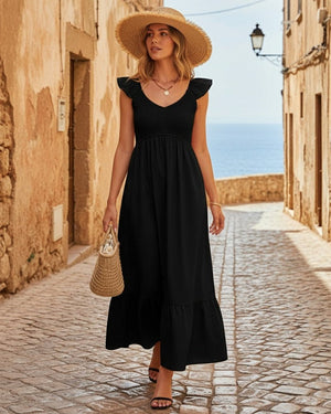 Woman in a black dress and straw hat standing in a sunlit alley with ocean view.