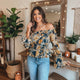 Woman wearing a floral off-shoulder top and jeans in a home setting with plants and decor.