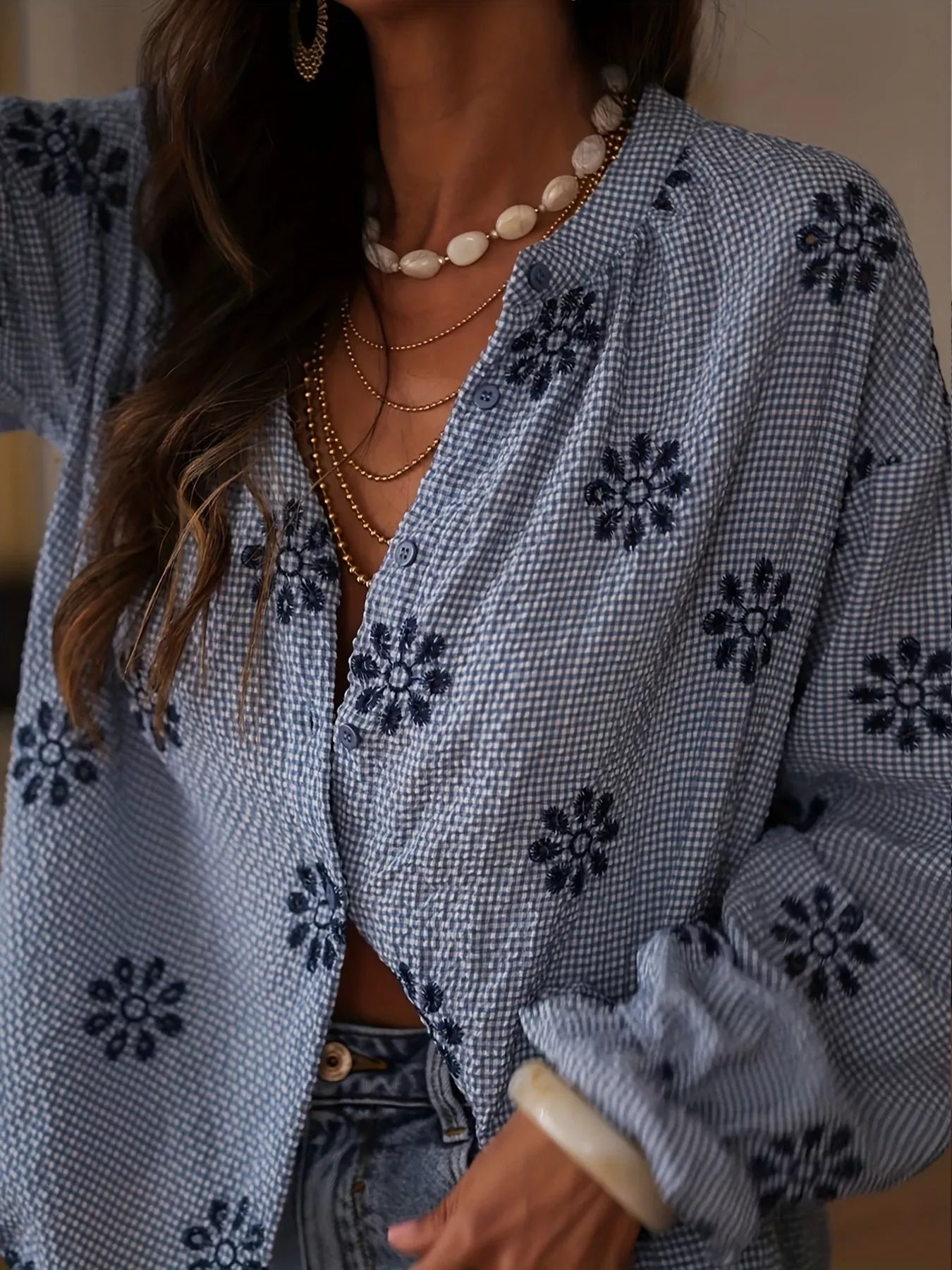 Person wearing a blue patterned cardigan with floral designs, layered necklaces, and denim jeans.