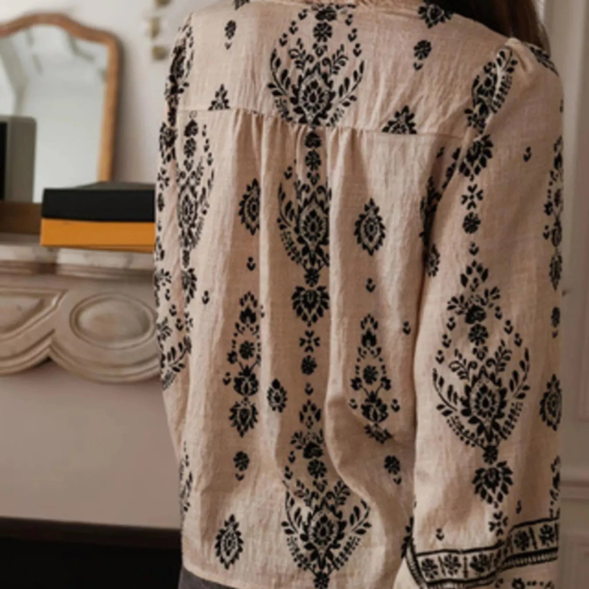 Beige blouse with black floral patterns worn by a person, with a blurred indoor background.