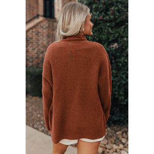 Person wearing a brown sweater standing outdoors with a brick wall and greenery in the background