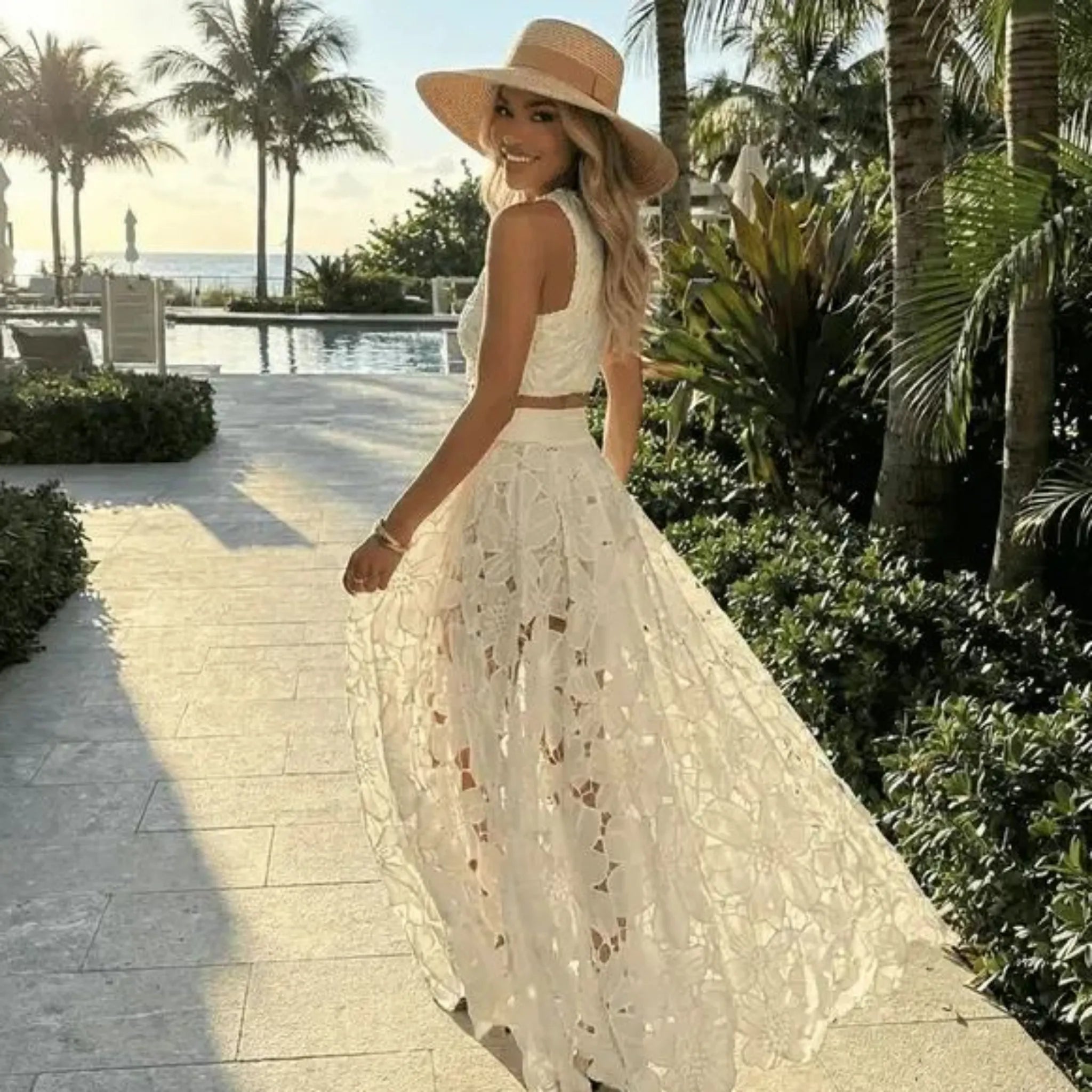 Woman in a white lace dress and straw hat walking along a path with palm trees and a pool in the background.