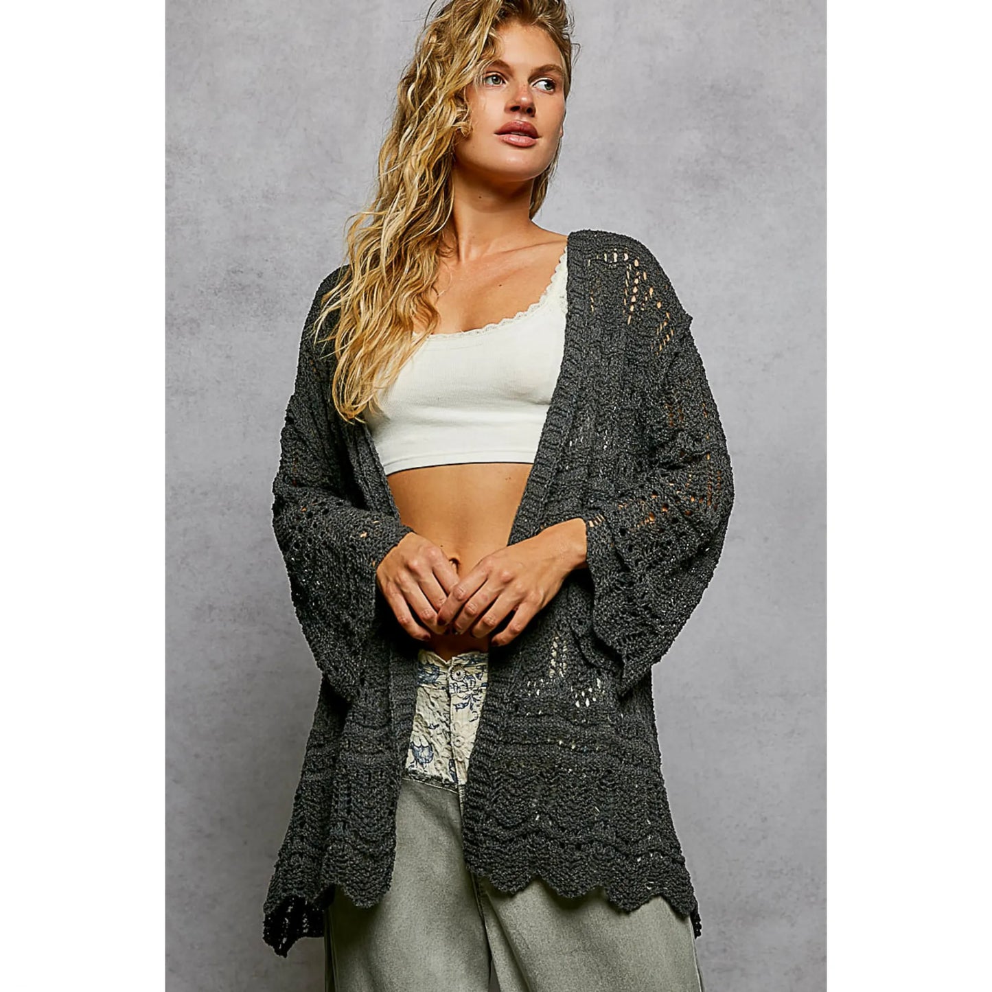 Woman wearing a dark gray crochet cardigan over a white top and light pants on a gray background