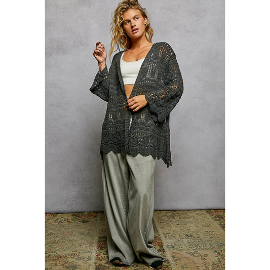 Woman wearing a dark lace cardigan over a white top and light green pants against a gray background.